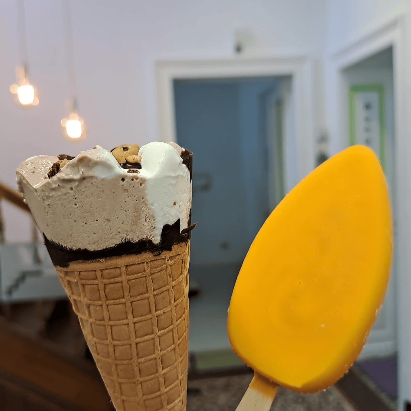 Zwei angebissene Eis vor dem Flur der Zahnarztpraxis: links ein Waffelhörnchen mit hellbrauner und weißer Eiscreme, Keksstückchen und dunkler Schokolade am Waffelrand, rechts ein gelbes Mango-Eis am Stiel. Im unscharfen Hintergrund zwei Hängeleuchten und die offenen Türen der Praxisräume.